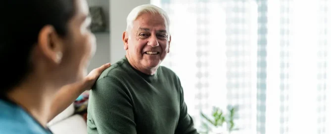 Senior man talking with a caregiver during a home medical visit, both calm and attentive at a table.